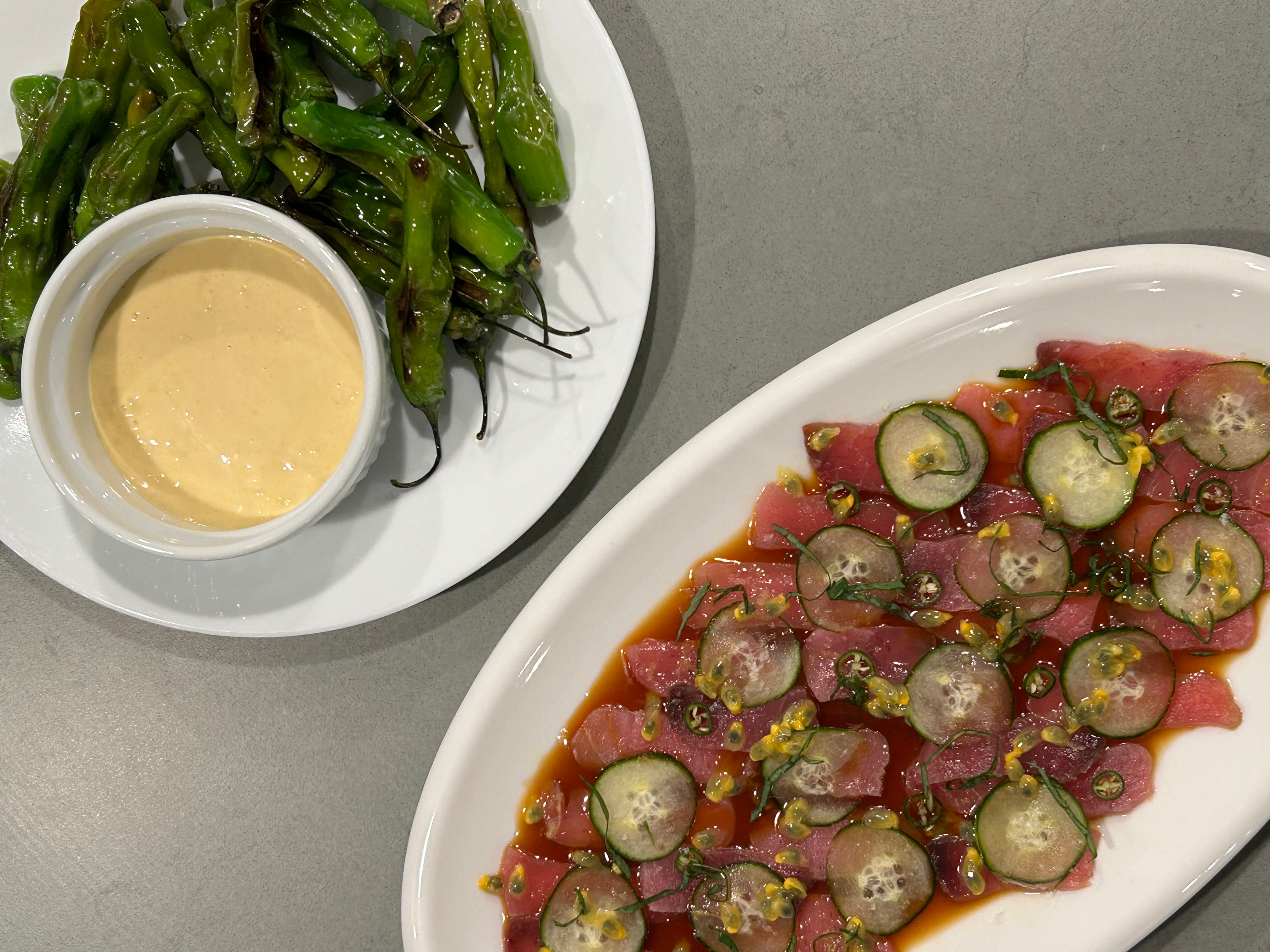 Plate of sliced raw fish with green olives and lemon zest, alongside a bowl of dipping sauce and green vegetables, on a gray surface.
