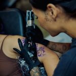 Tattoo artist with black gloves tattooing a purple design on a person's arm, woman with dark hair and face tattoo, focused.