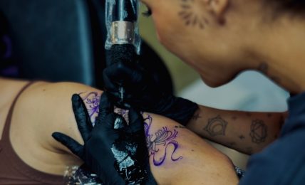 Tattoo artist with black gloves tattooing a purple design on a person's arm, woman with dark hair and face tattoo, focused.