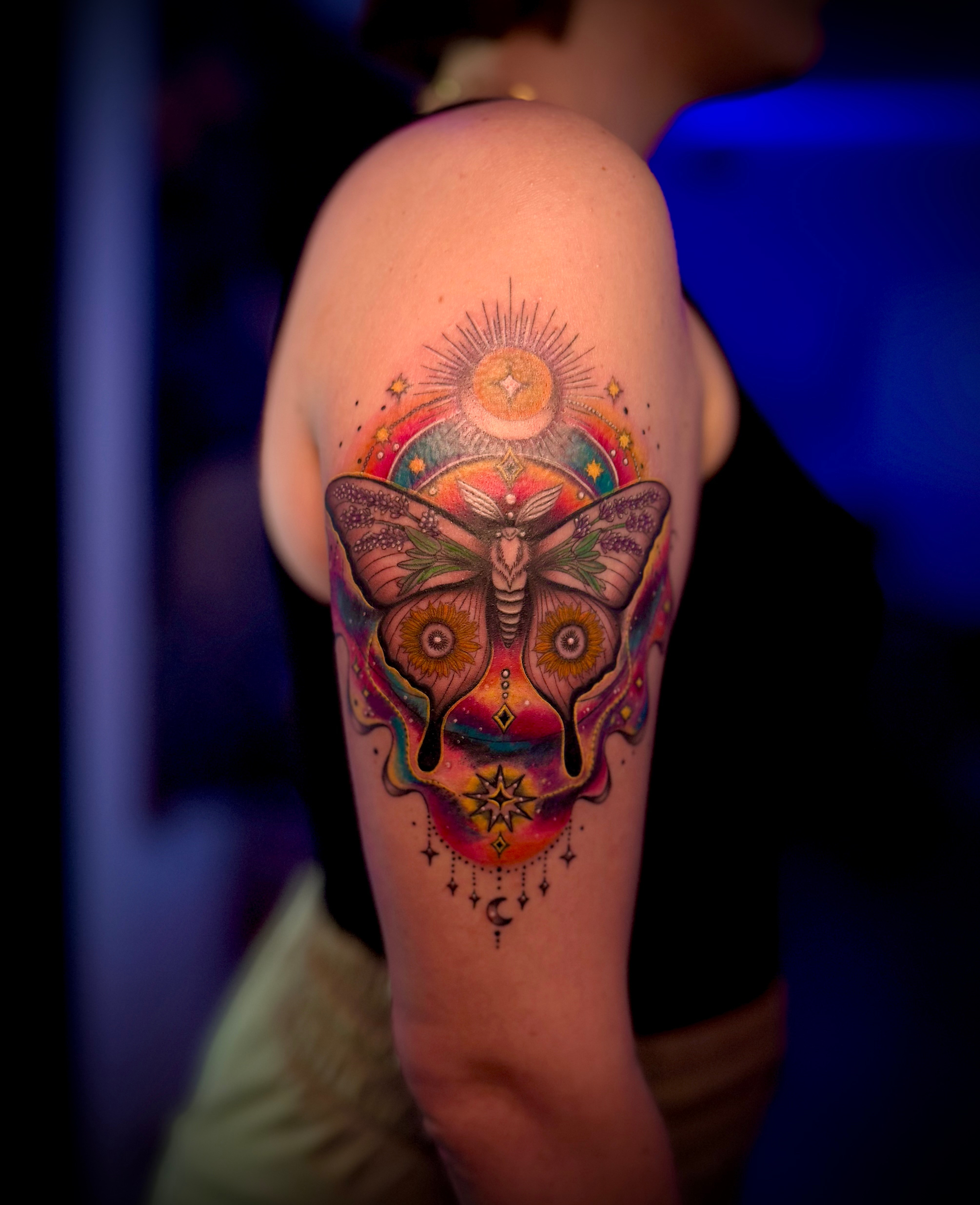 Colorful tattoo of a butterfly with geometric patterns on upper arm, surrounded by rainbow and sun design, against dark background.