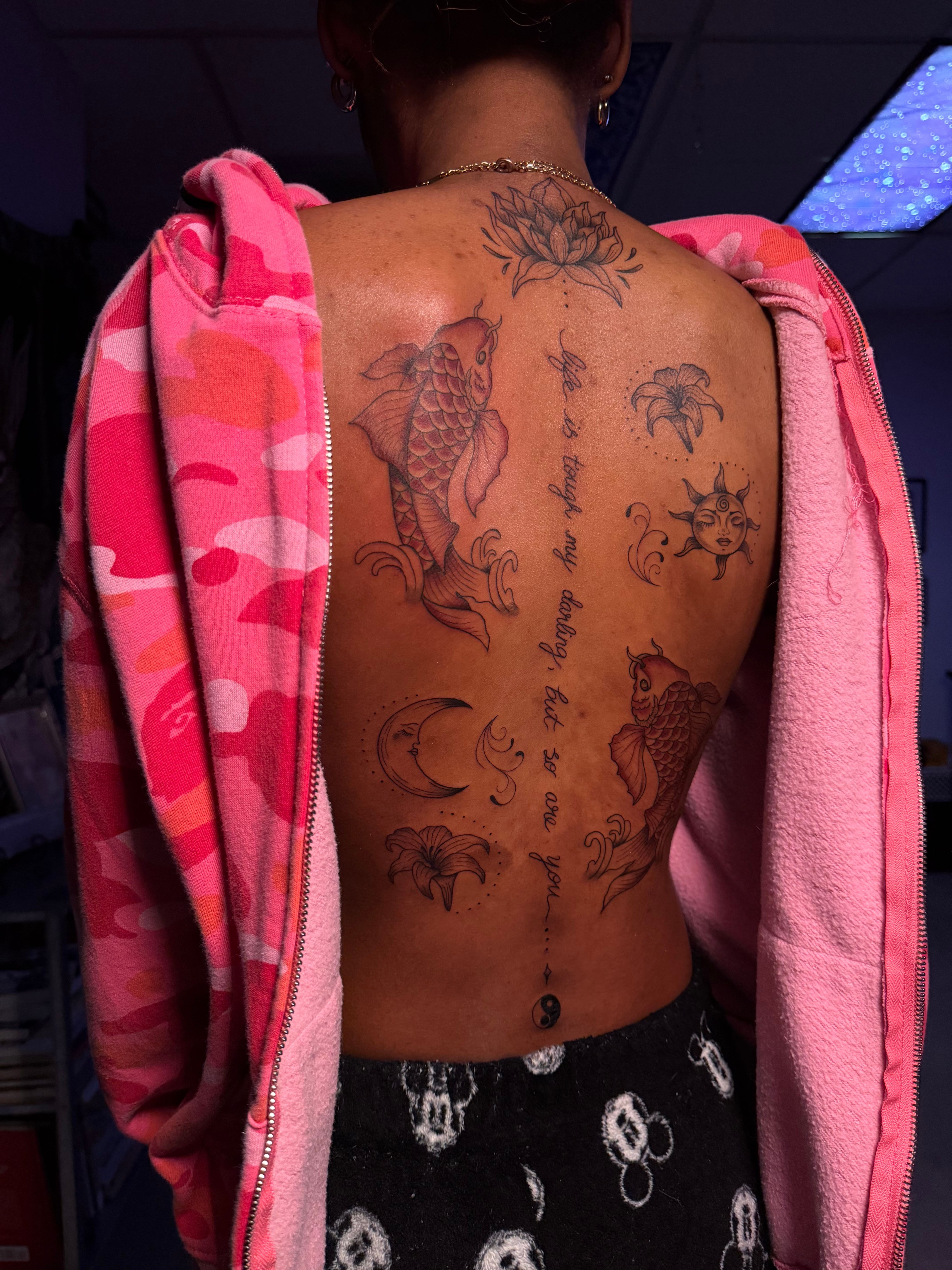 Person's back with tattoos, wearing a pink hoodie and black pants with white patterns, lying down.