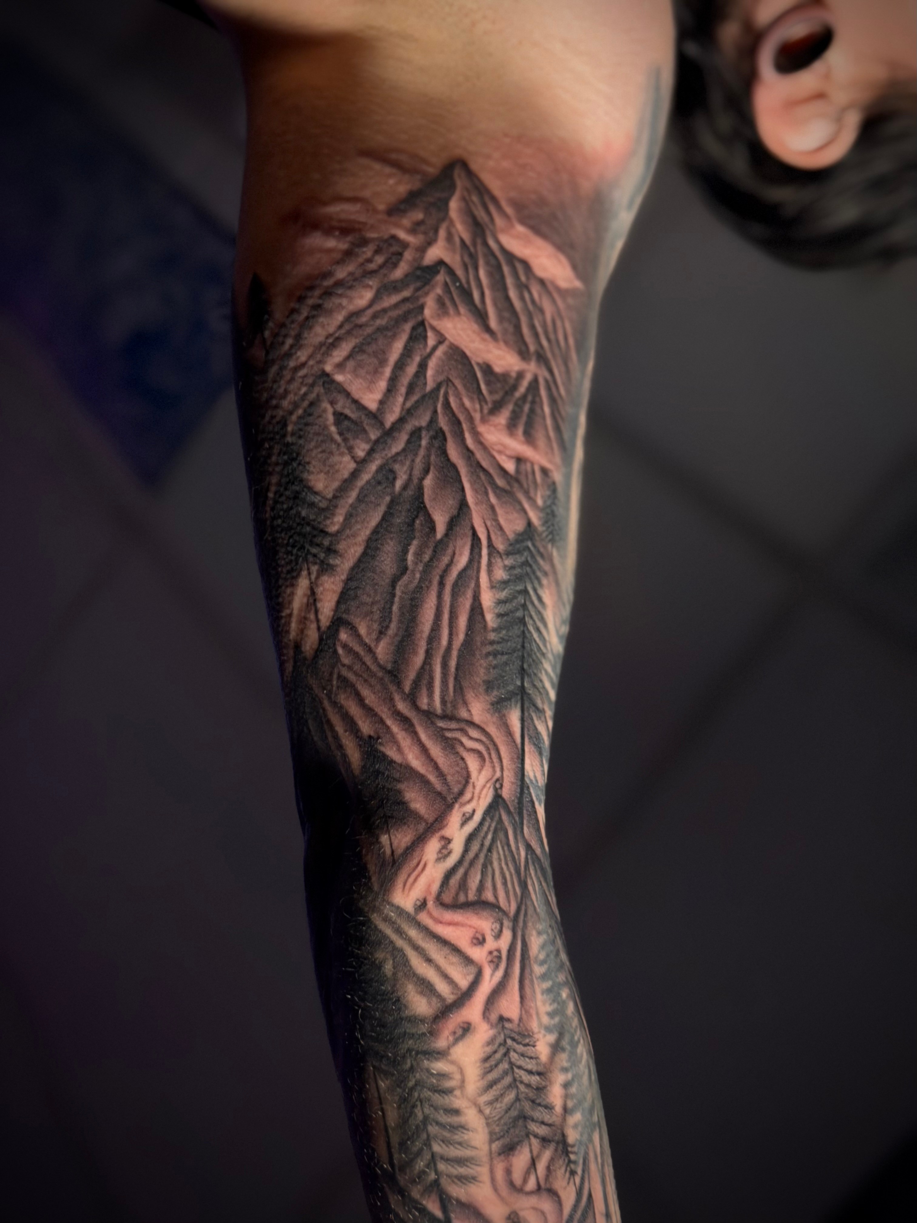 Tattoo of mountain peaks, trees, and a winding path on a person's arm, detailed and shaded.