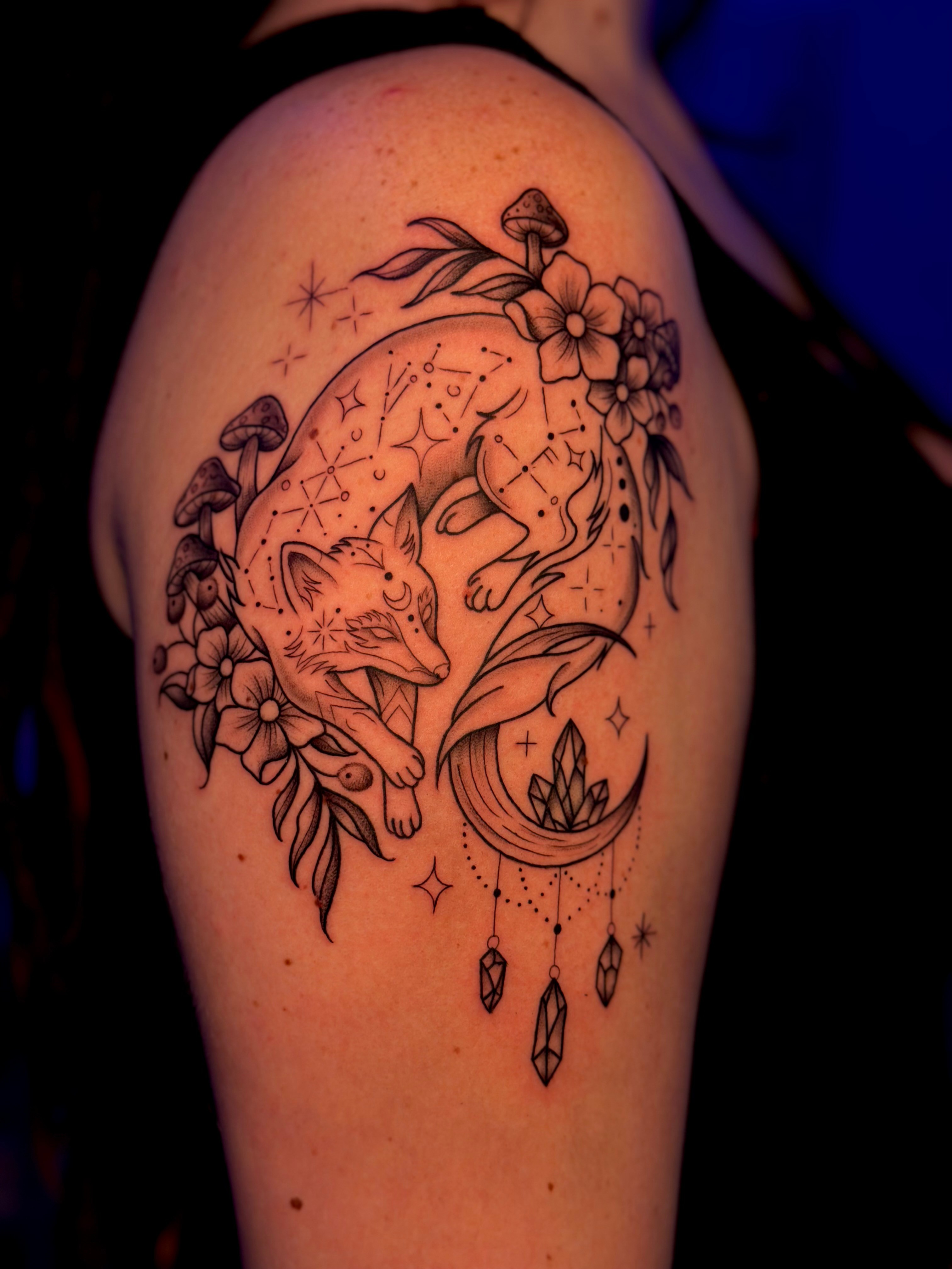Tattoo of a fox surrounded by flowers, a crescent moon, and hanging crystals on upper arm.