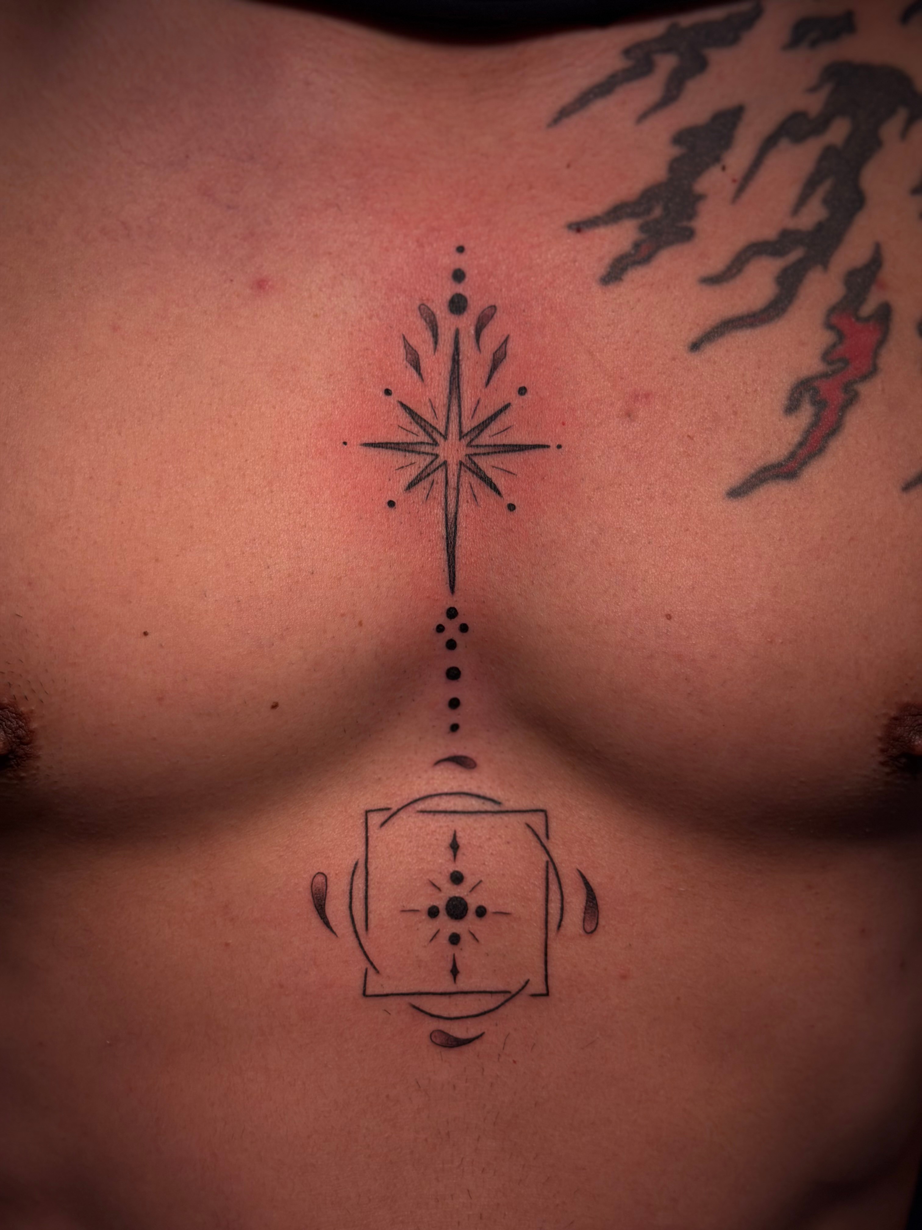 Tattoo of a compass star with dotted line above a stylized face on chest, with additional tattoos on shoulder. Word count: 20