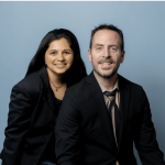 Two people, a woman and a man, pose together against a light blue background, smiling at the camera.