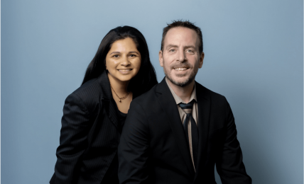 Two people, a woman and a man, pose together against a light blue background, smiling at the camera.