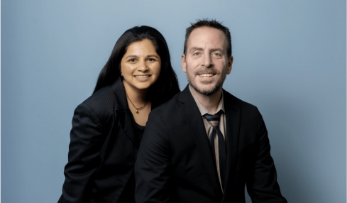 Two people, a woman and a man, pose together against a light blue background, smiling at the camera.