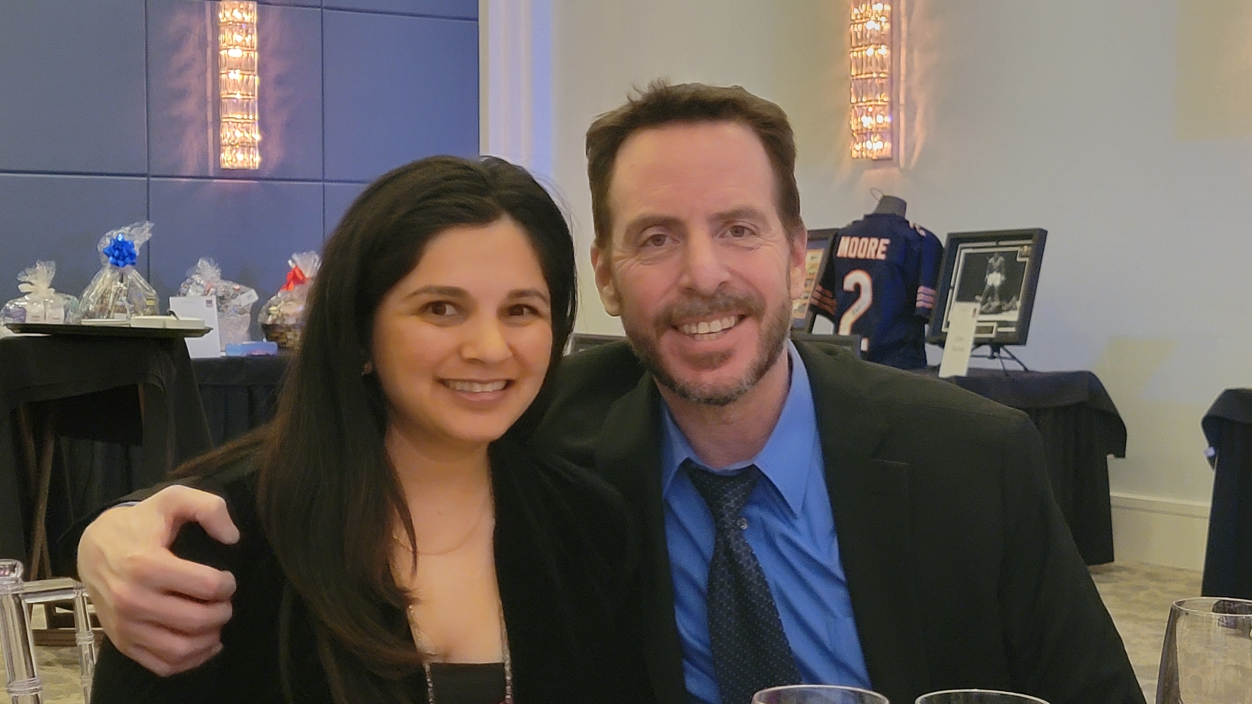Two people smiling and posing together at an indoor event, with a table and framed photo in the background.