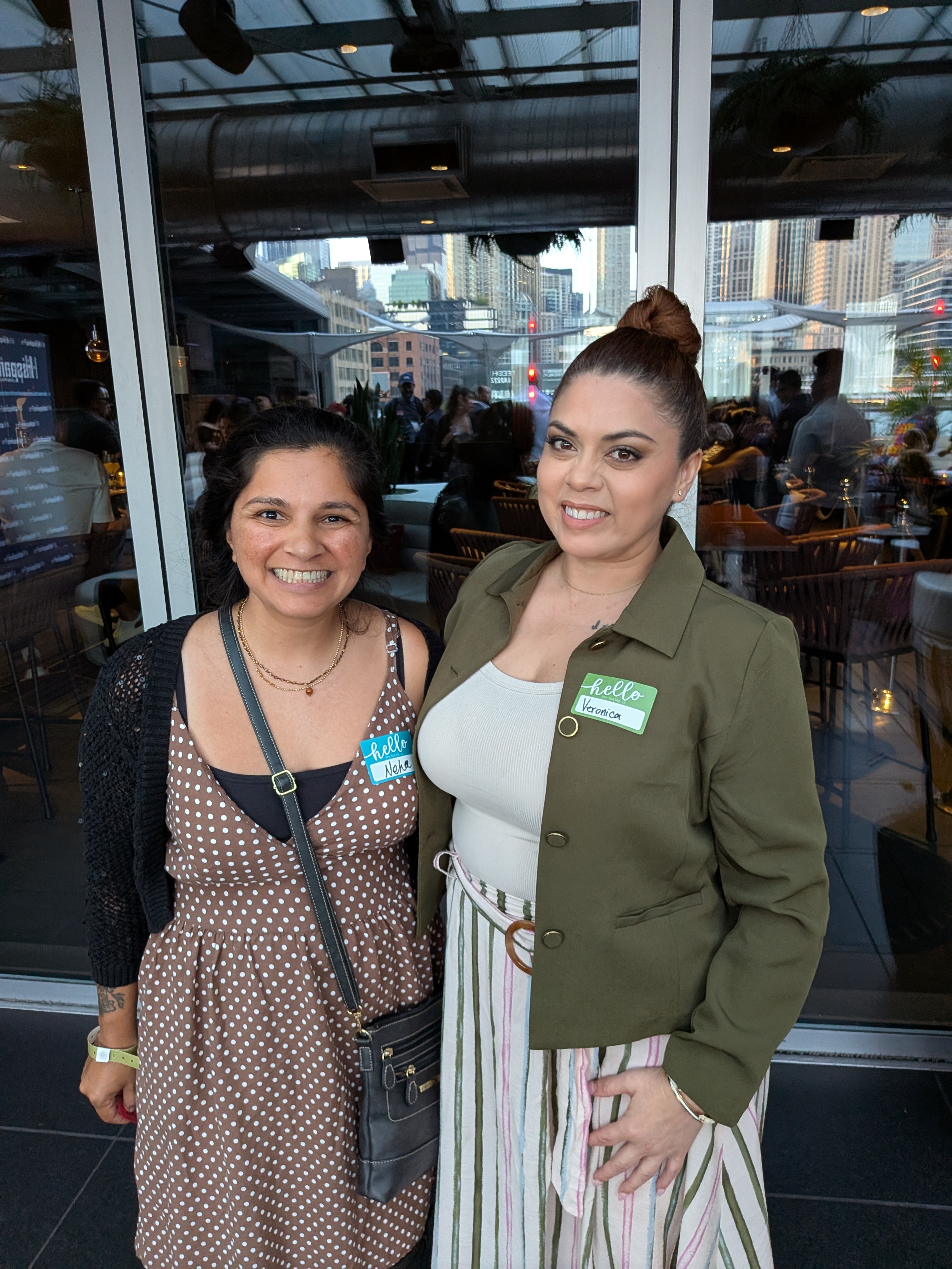 Two women smiling indoors with cityscape visible through large windows behind them.