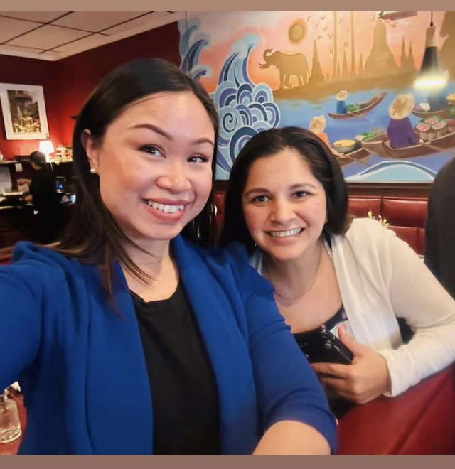 Two women smiling in a restaurant with colorful mural background, one in blue jacket, the other in white, holding a phone.