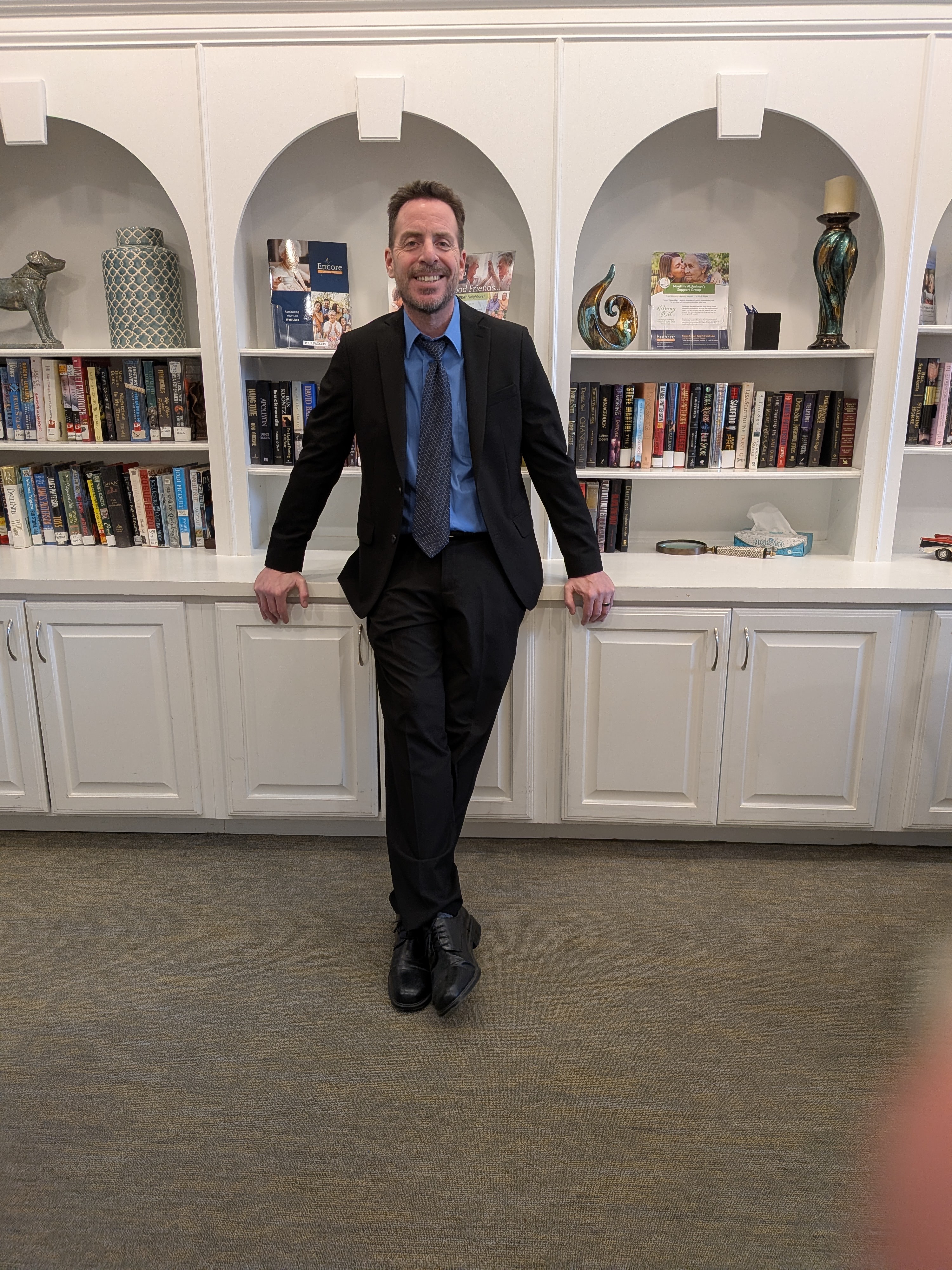 Man in a suit standing in front of a white bookshelf with decorative items and books, smiling.