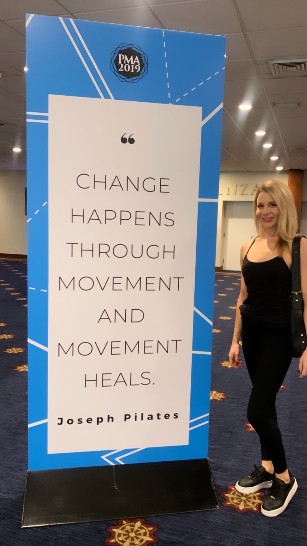 Standing sign with quote about change, movement, and healing, attributed to Joseph Pilates, in a room with a woman nearby.
