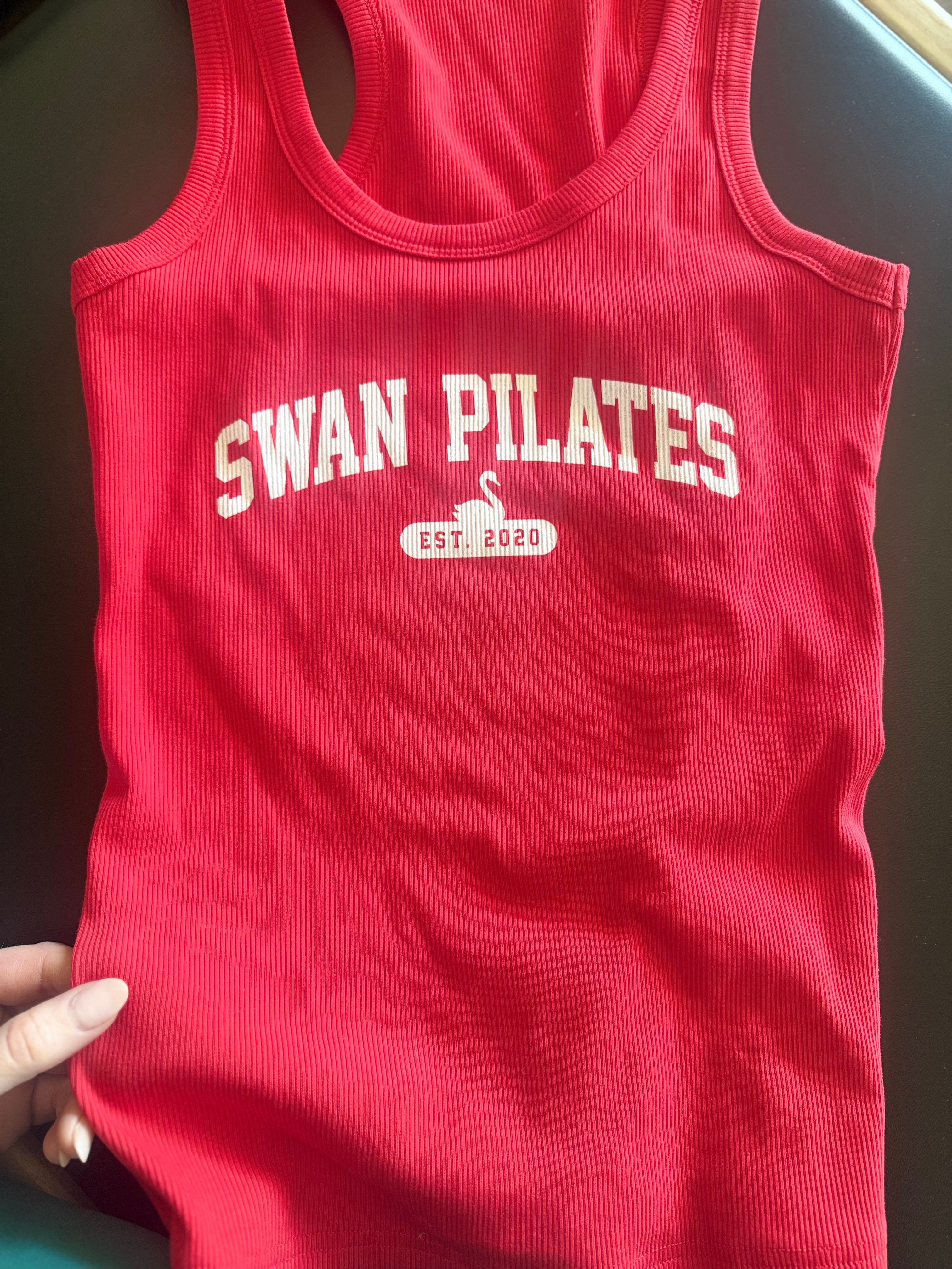 Red tank top with white text reading 'SWAN PILATES' and a swan logo with 'EST. 2020' beneath.