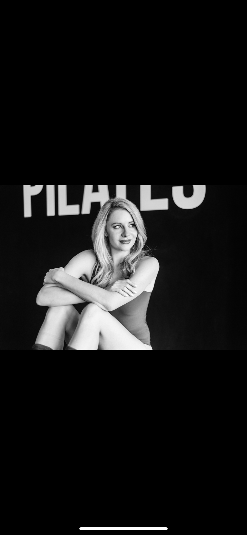 Young woman sitting with crossed legs in front of a black background with large text, smiling slightly.