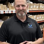 Man with short hair and beard wearing a black polo shirt with a logo, standing in a warehouse with shelves of boxes.