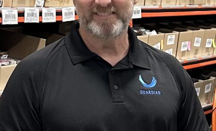 Man with short hair and beard wearing a black polo shirt with a logo, standing in a warehouse with shelves of boxes.