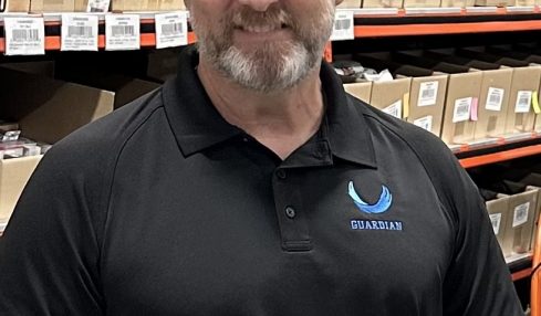 Man with short hair and beard wearing a black polo shirt with a logo, standing in a warehouse with shelves of boxes.