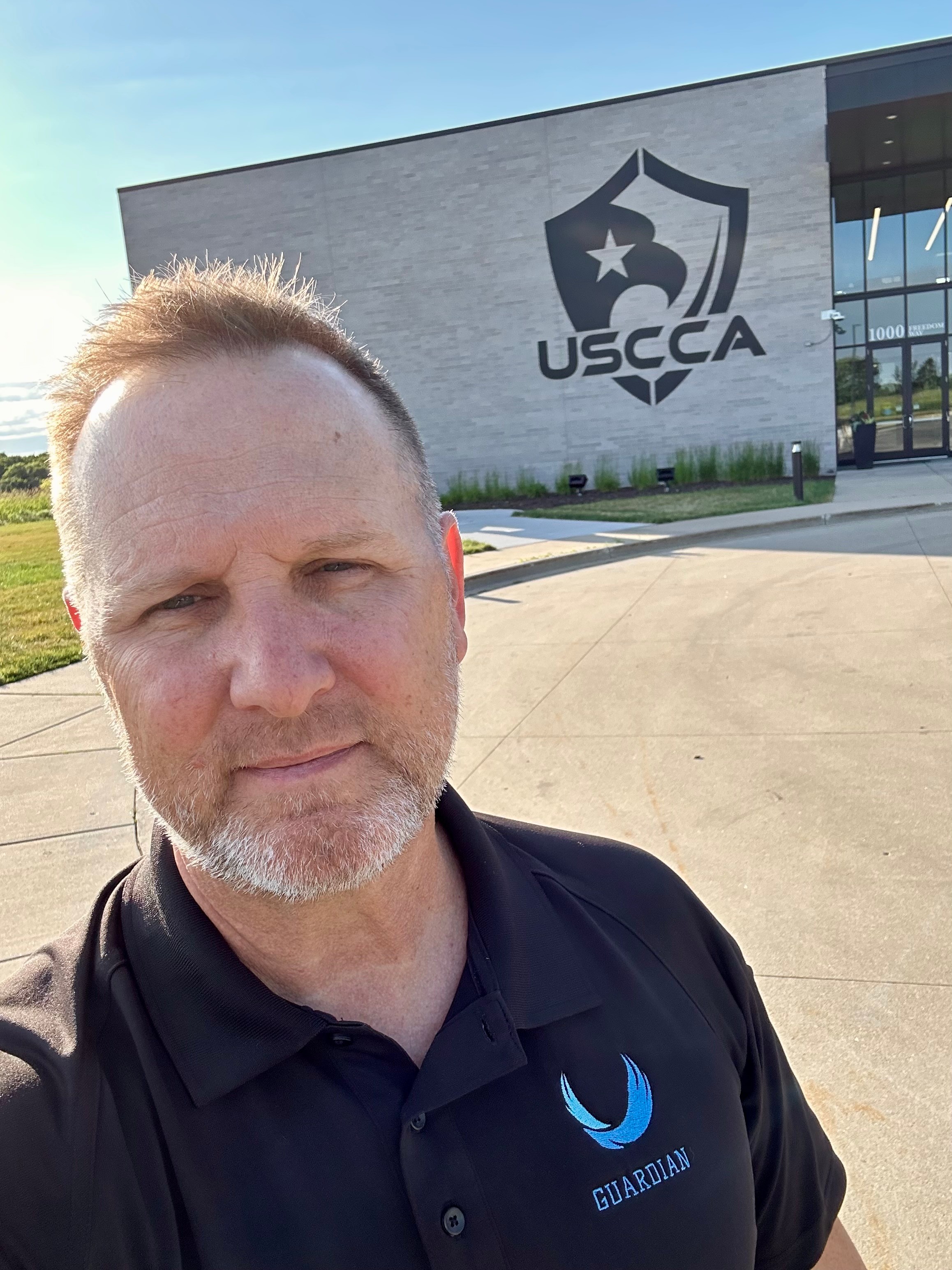 Man with short hair and beard taking selfie outside USCCA building, wearing a black shirt with a blue logo.