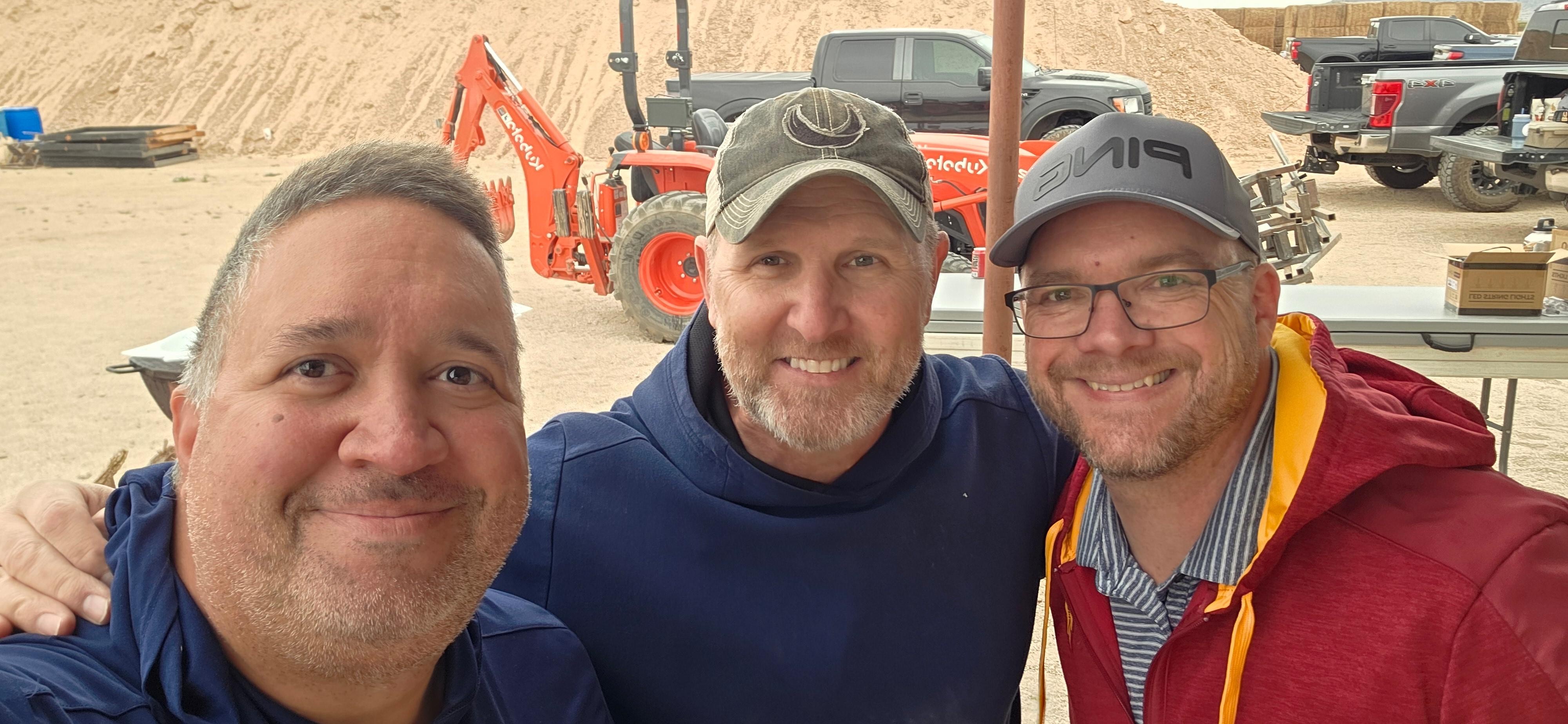 Three men smiling outdoors, with construction equipment and vehicles in the background.