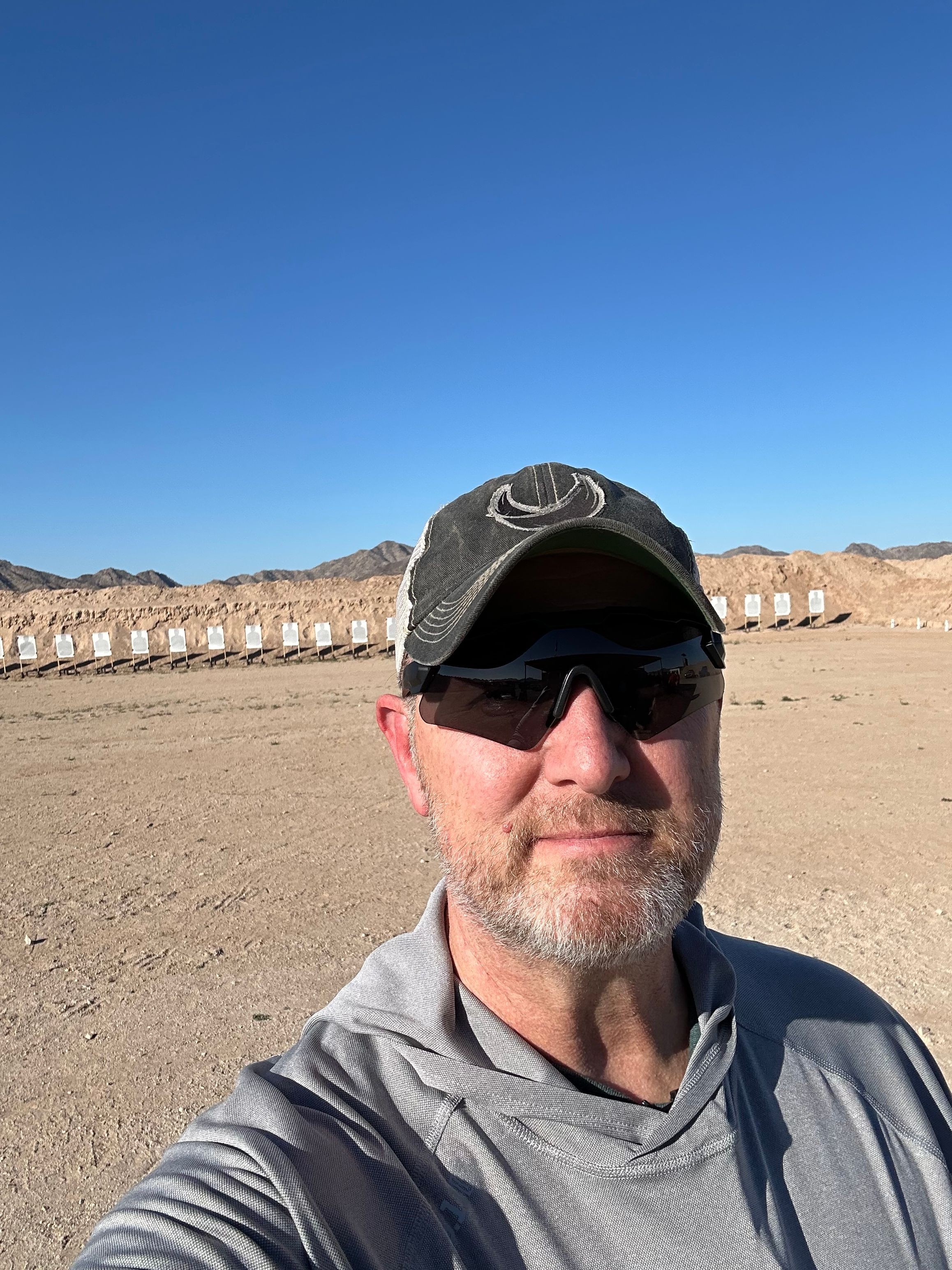 Man wearing sunglasses and a cap takes a selfie on a sandy beach with a clear blue sky.