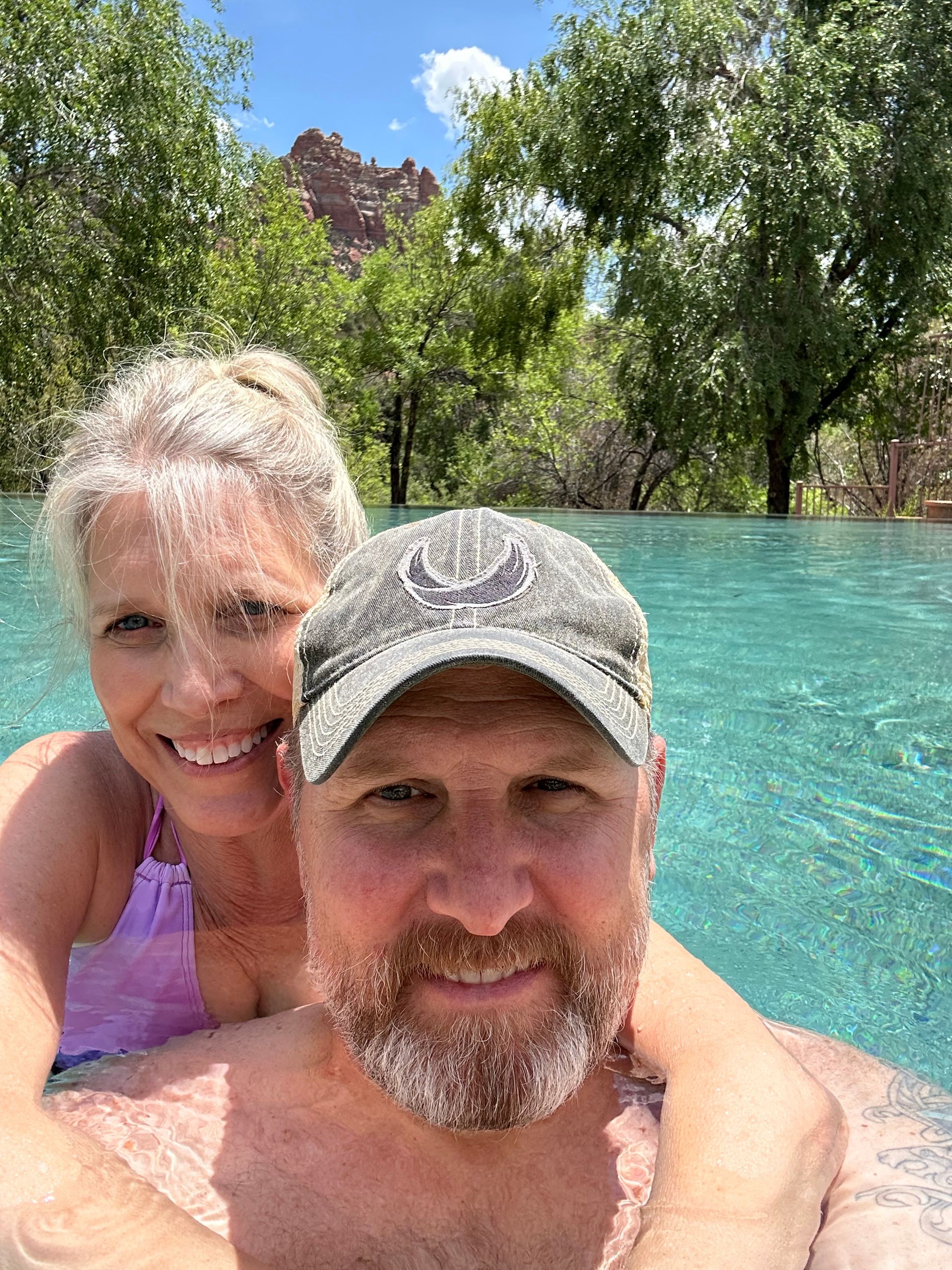 Two people swimming in a pool, smiling, with trees and blue sky in the background.