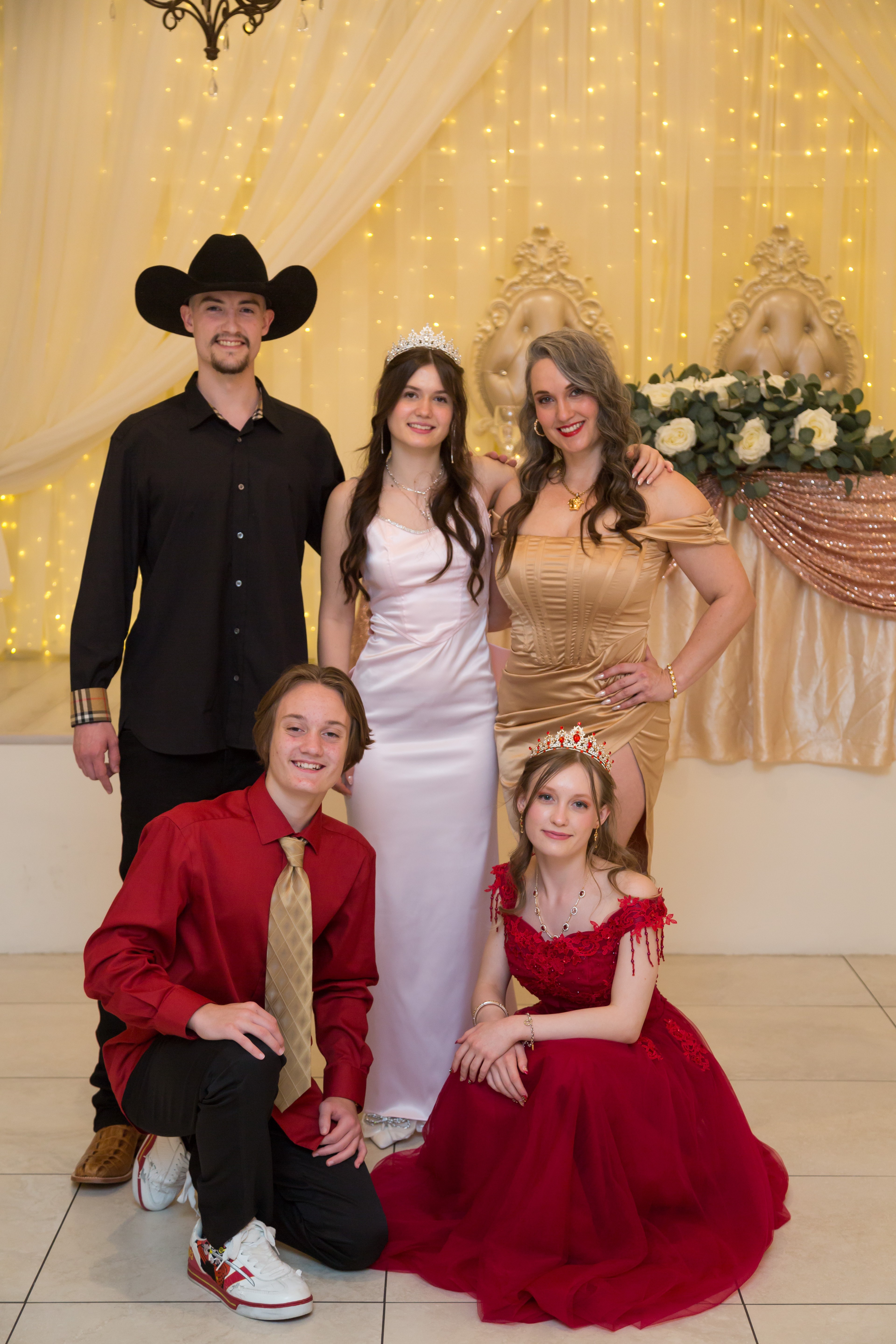 Group of five people dressed in formal attire, standing in front of decorated backdrop with lights and flowers.