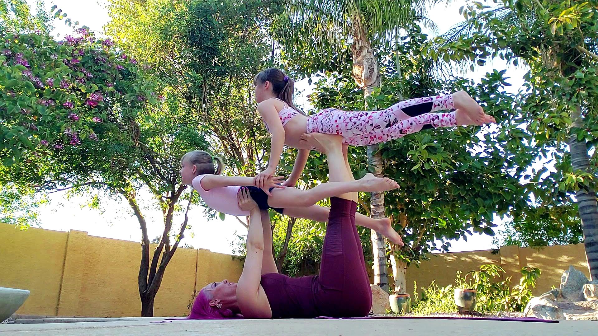 Two women practicing acro yoga outdoors with trees and a yellow wall in the background.