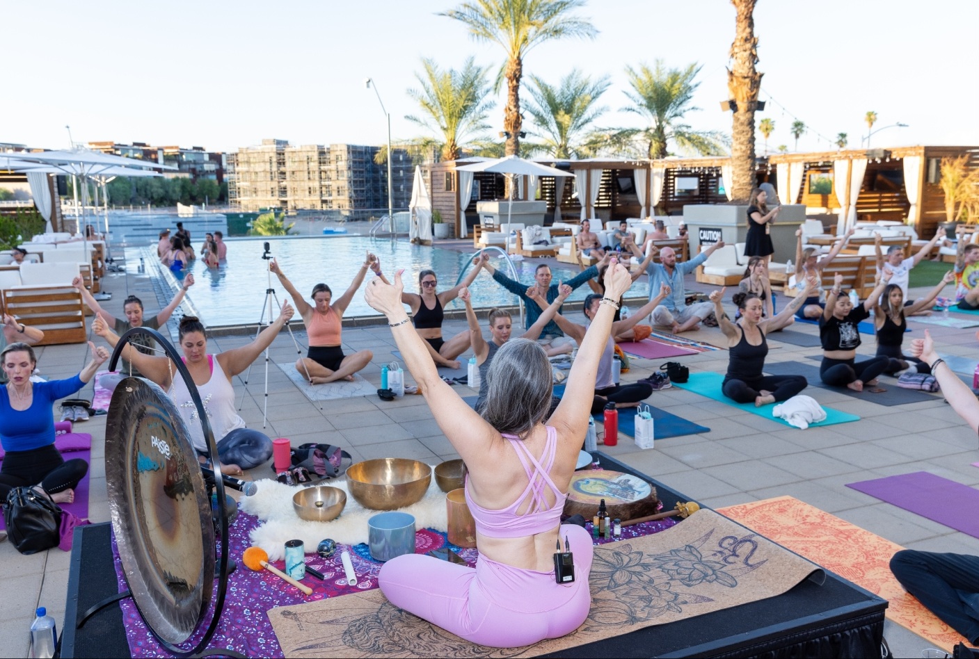 Group of people practicing yoga outdoors near palm trees with a cityscape in the background, seated on mats with arms raised.