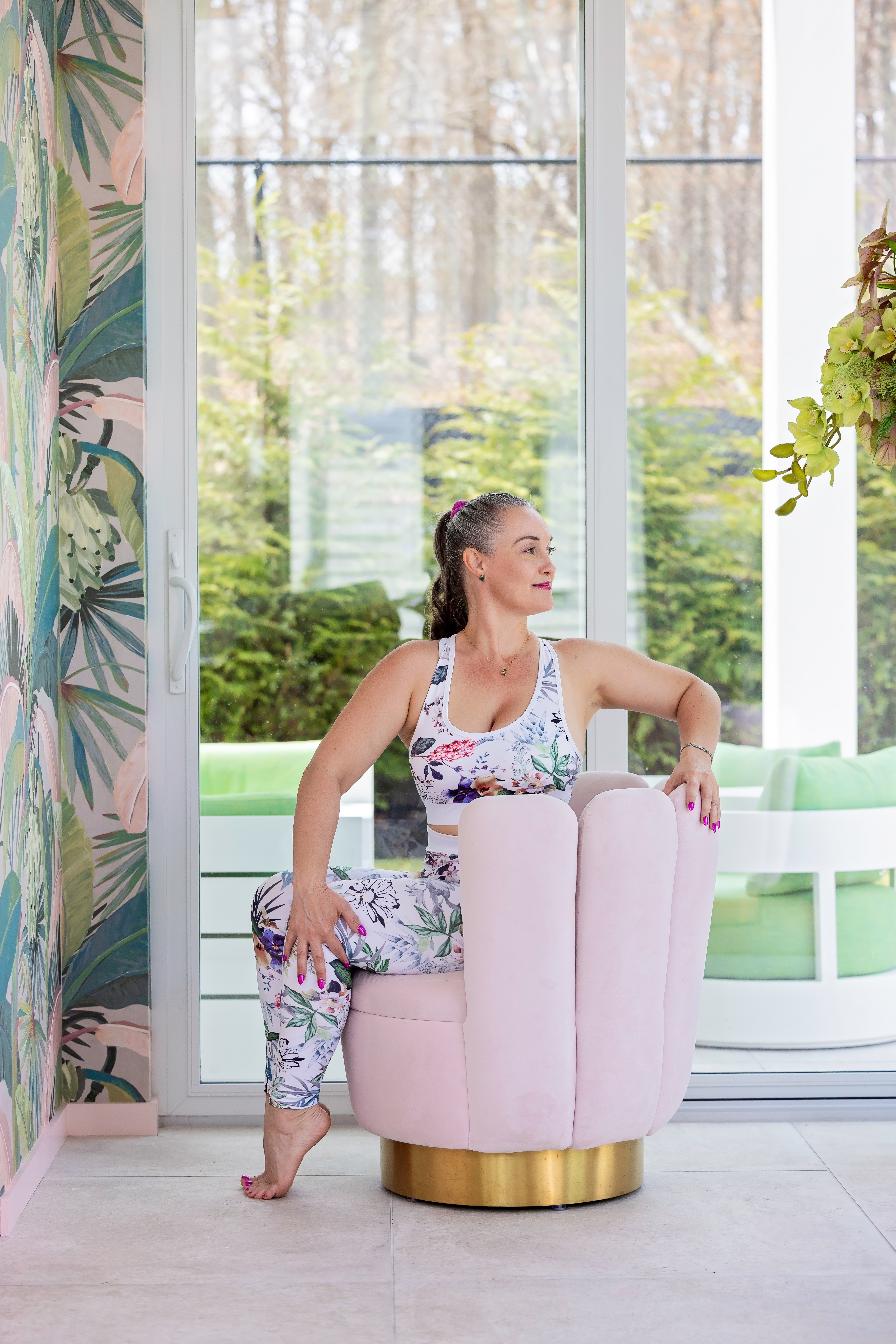 Woman in floral workout clothes sitting on a pink armchair near large window with greenery outside.