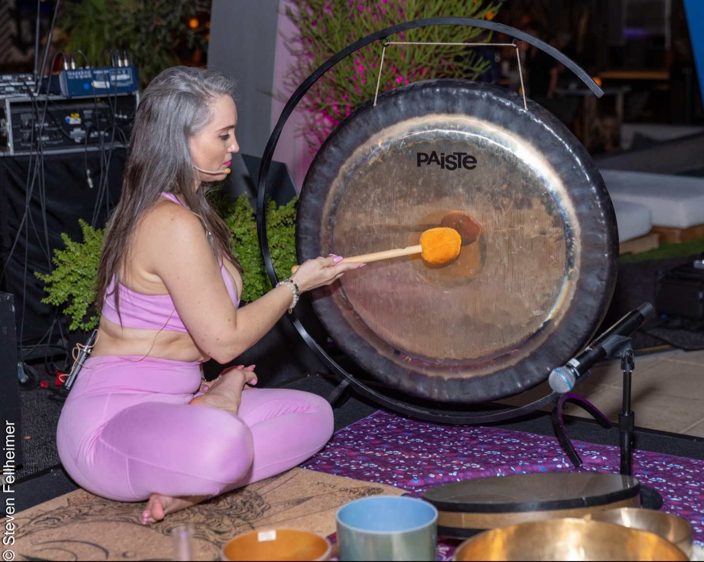 Young woman in pink clothing playing a large gong with a mallet, seated on a patterned mat indoors.