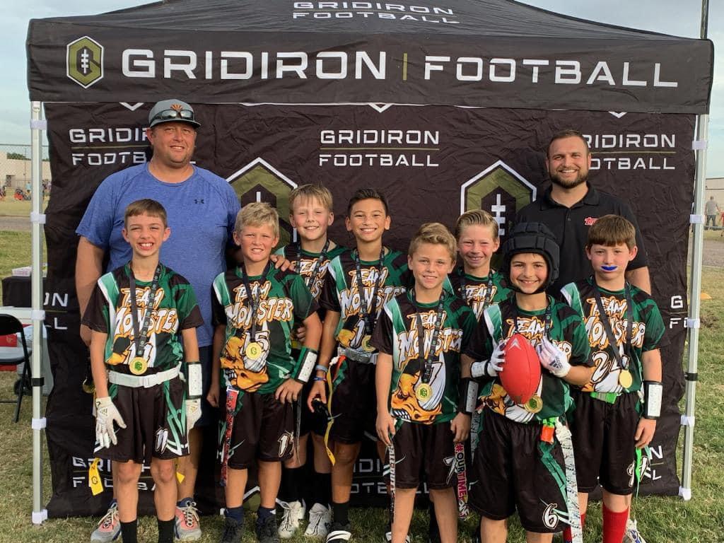 Group of young football players with medals and a coach standing in front of a black tent with 'Gridiron Football' text.