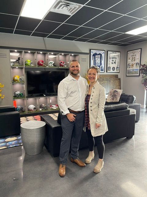 Two people standing in a modern office lounge area with shelves, a TV, and seating, one wearing a white coat and the other in a suit.