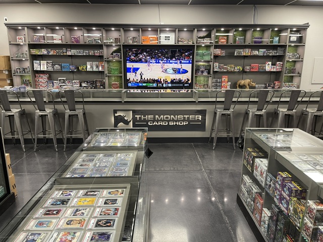 Video game store with shelves of games, a TV screen showing a sports event, and chairs in front of a counter.