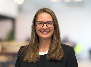 Woman with glasses smiling, wearing a black blazer, in an indoor setting with blurred background.