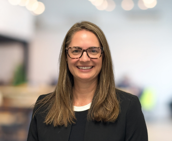 Woman with glasses smiling, wearing a black blazer, in an indoor setting with blurred background.