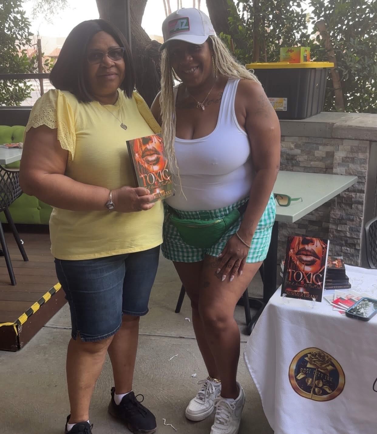 Two women standing outdoors, one holding a book, smiling, with trees and a table nearby.
