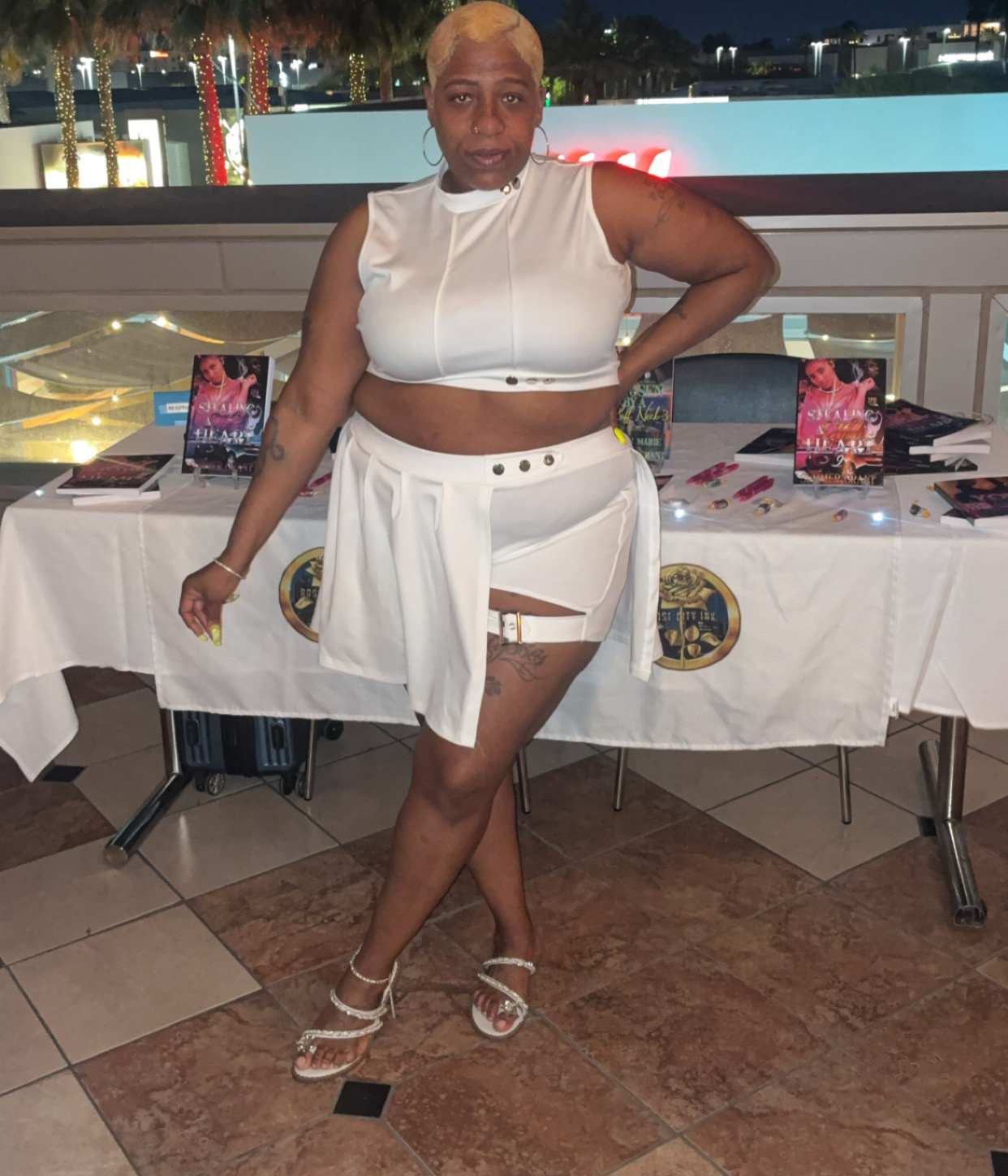 Woman with short blonde hair in white outfit, standing indoors with tables and promotional materials behind her.