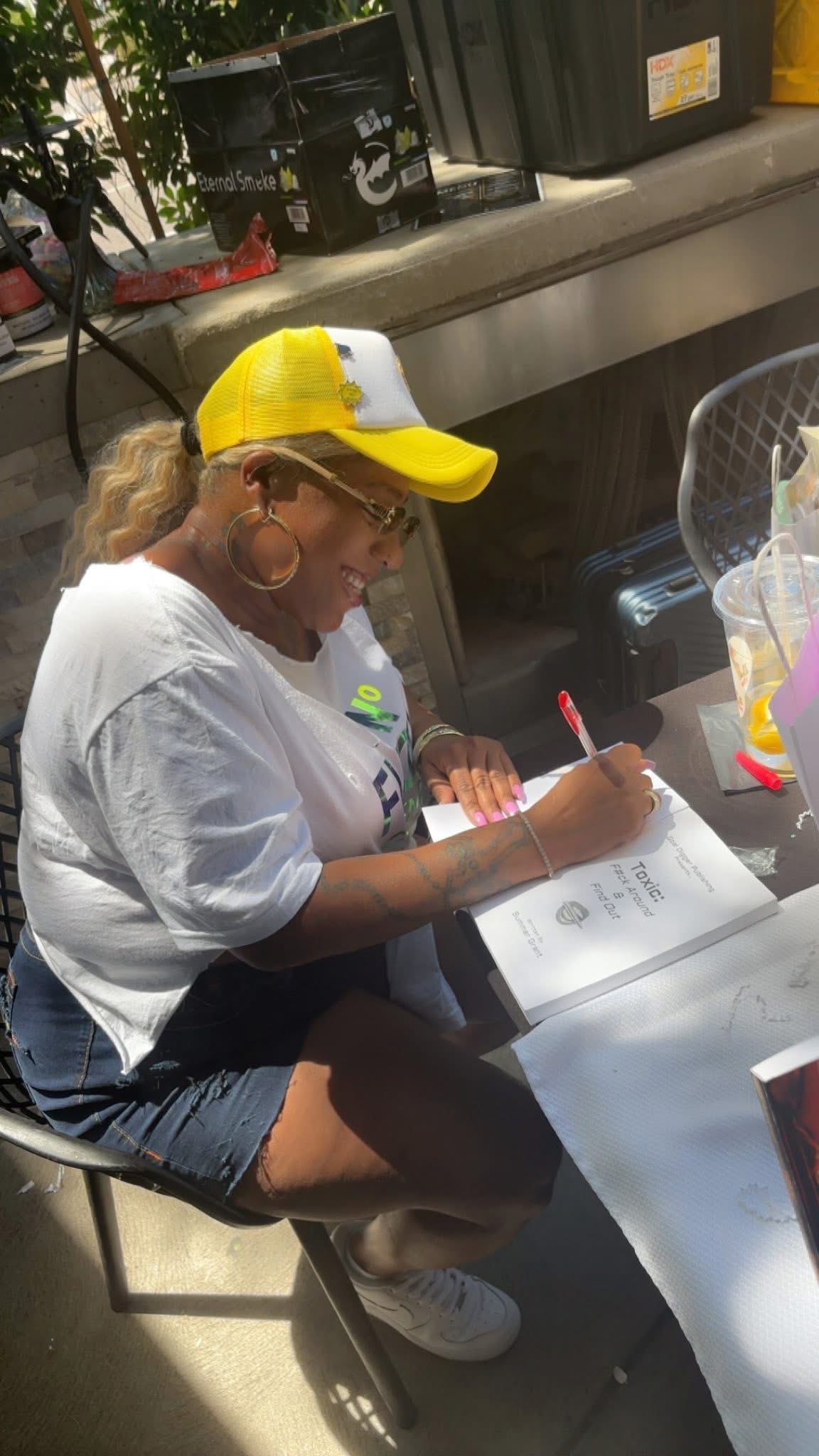 Woman wearing a yellow cap and white shirt sitting at a table, writing on paper, with a plastic cup nearby.