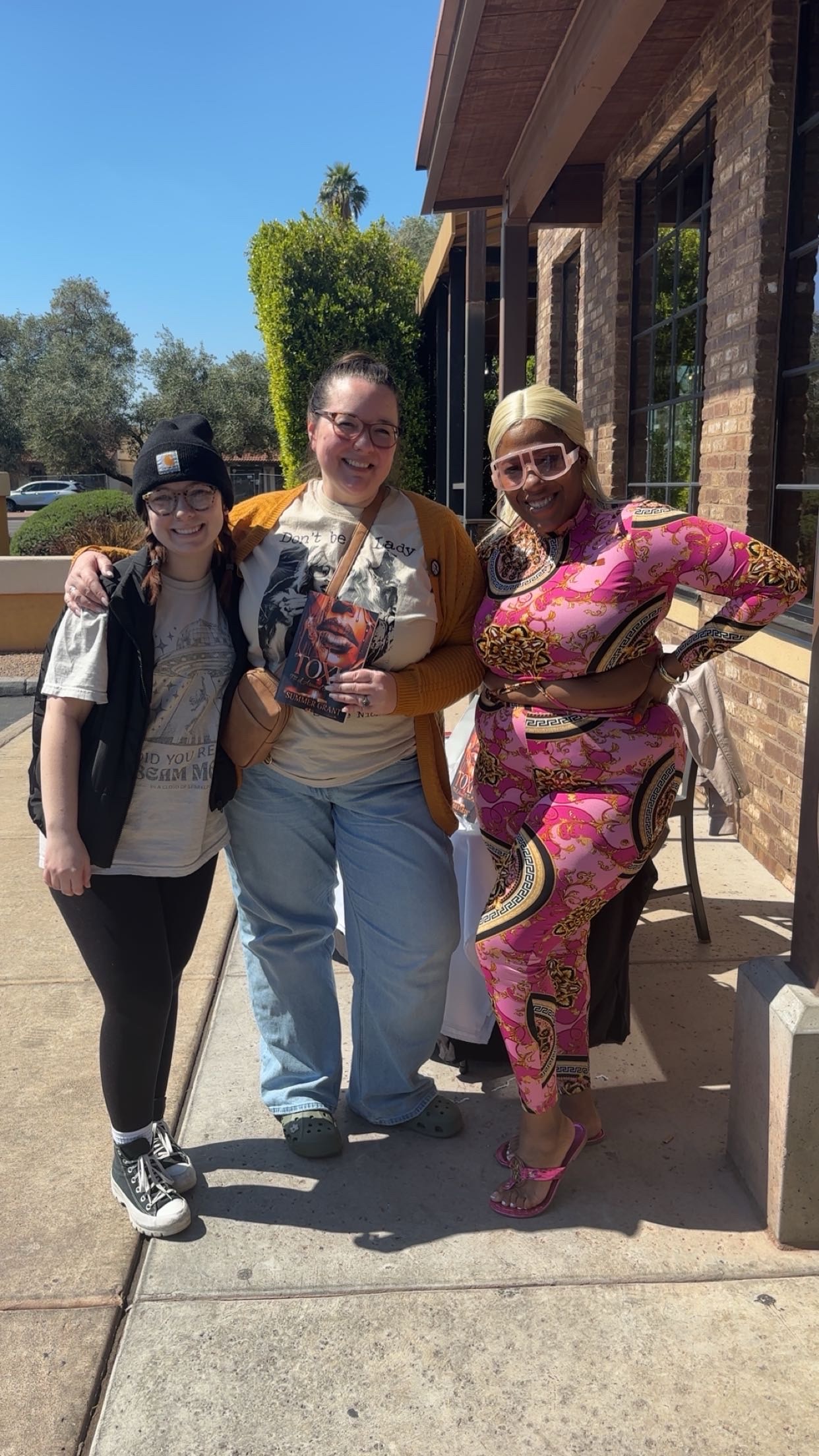 Three women standing outdoors, smiling, with trees and a building in the background.