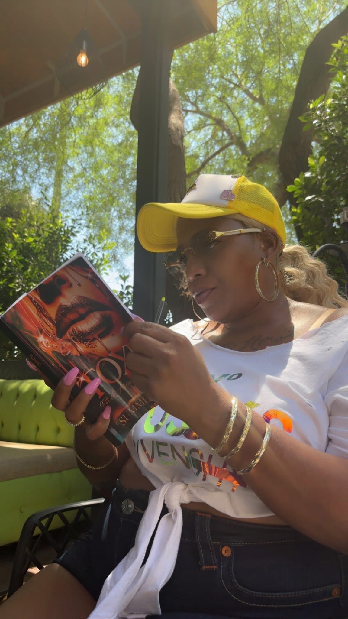 Woman wearing a yellow cap and sunglasses reads a magazine outdoors under trees.