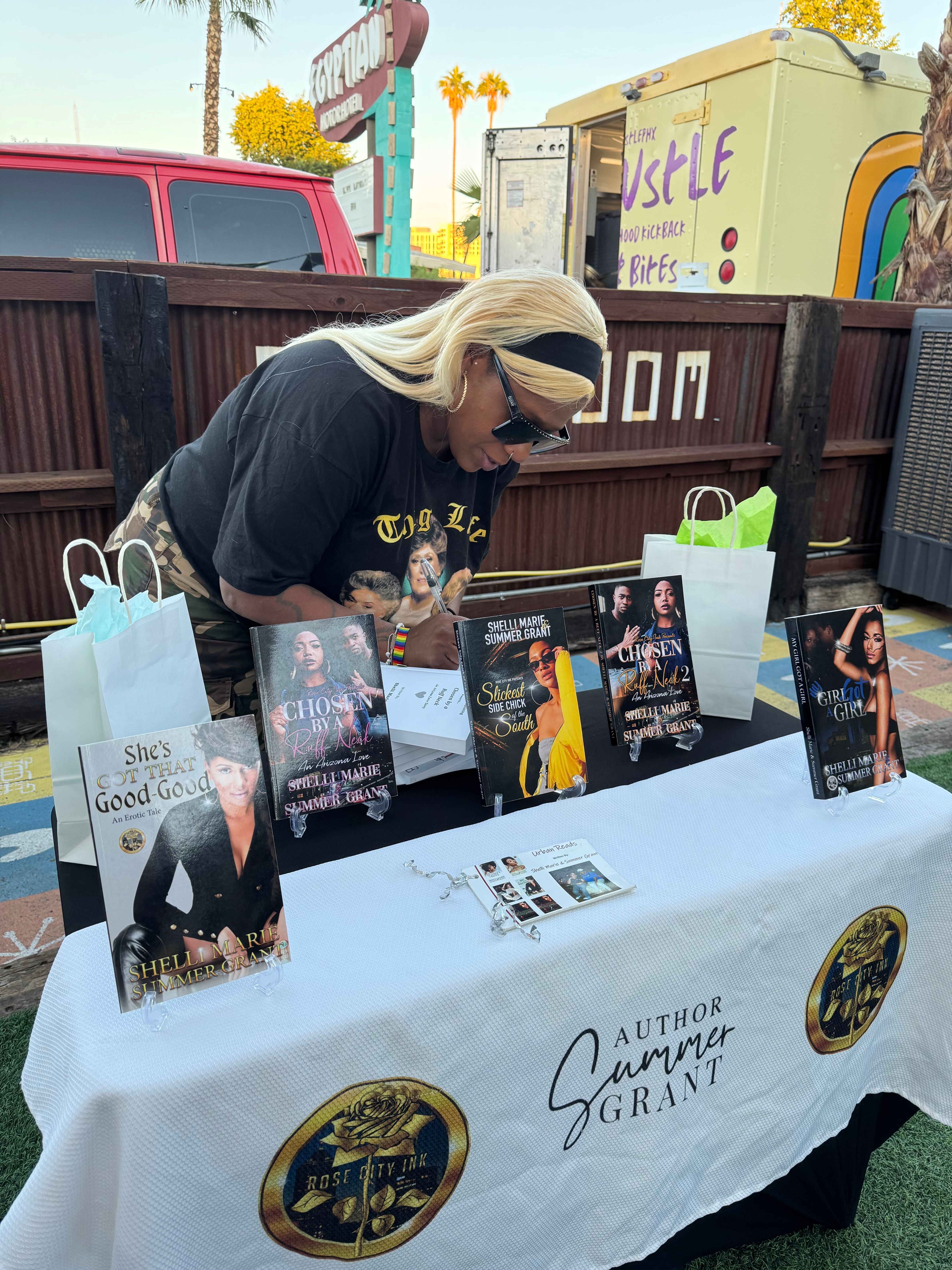 Woman with blonde hair and sunglasses sitting at a table with books and promotional materials outdoors.