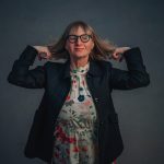 Woman with glasses and shoulder-length hair, wearing a floral dress and dark jacket, poses with hands behind her head against a dark background.