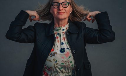 Woman with glasses and shoulder-length hair, wearing a floral dress and dark jacket, poses with hands behind her head against a dark background.