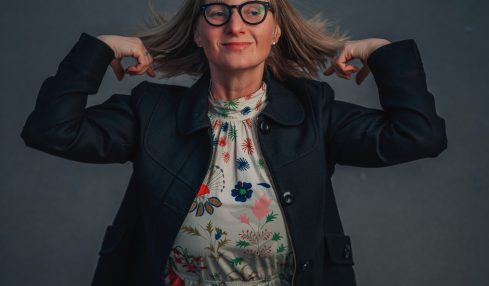Woman with glasses and shoulder-length hair, wearing a floral dress and dark jacket, poses with hands behind her head against a dark background.