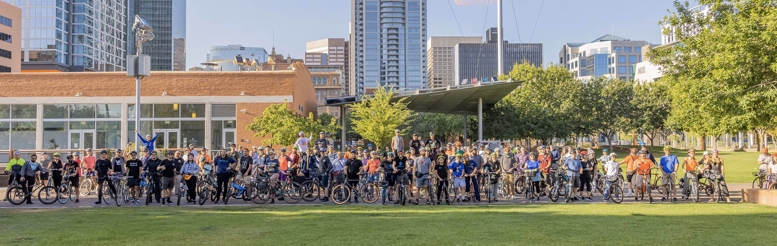 Large group of people gathered outdoors in a park with city buildings in the background.