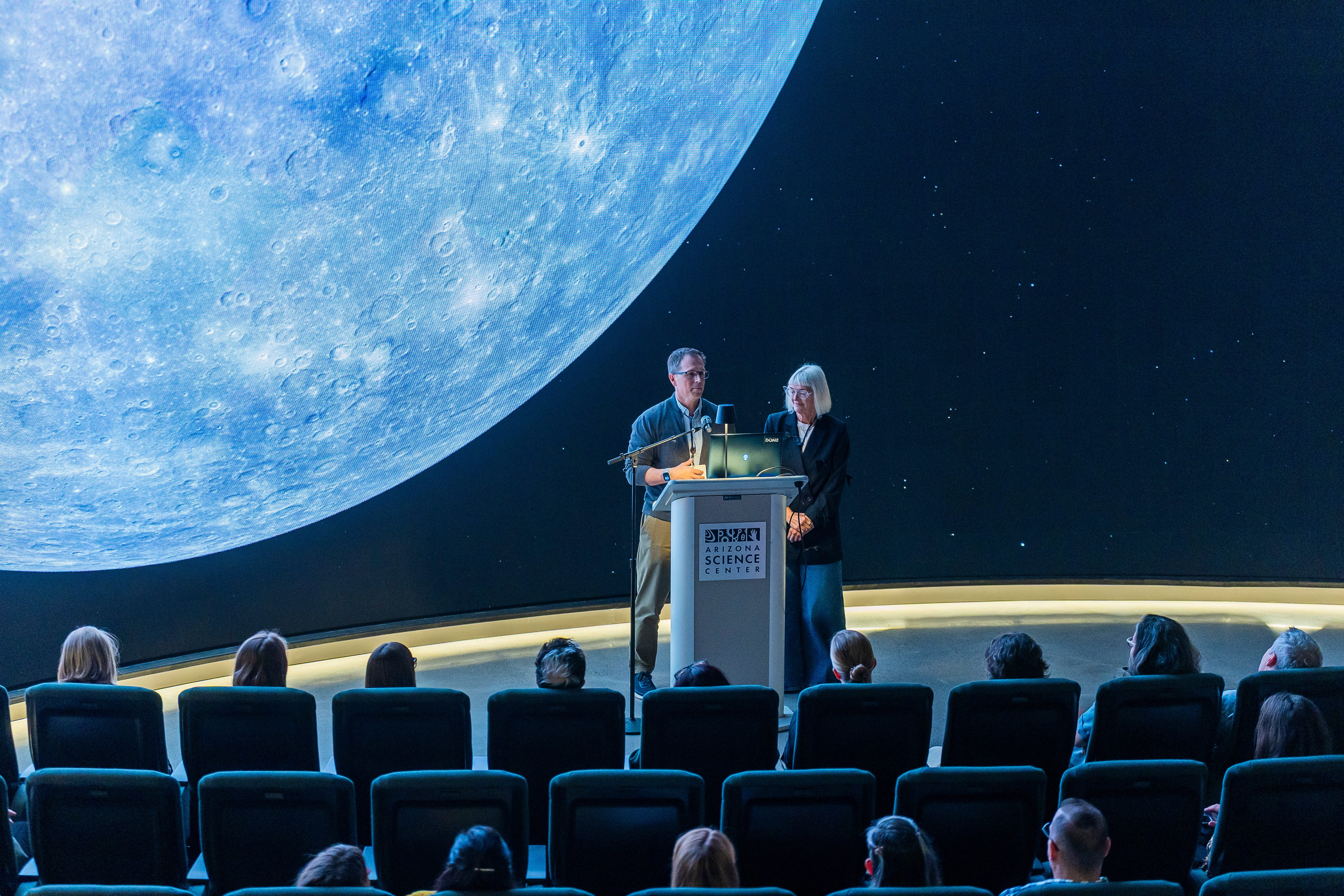 Two people stand at a podium on stage with a large moon projection behind them, audience seated in front.
