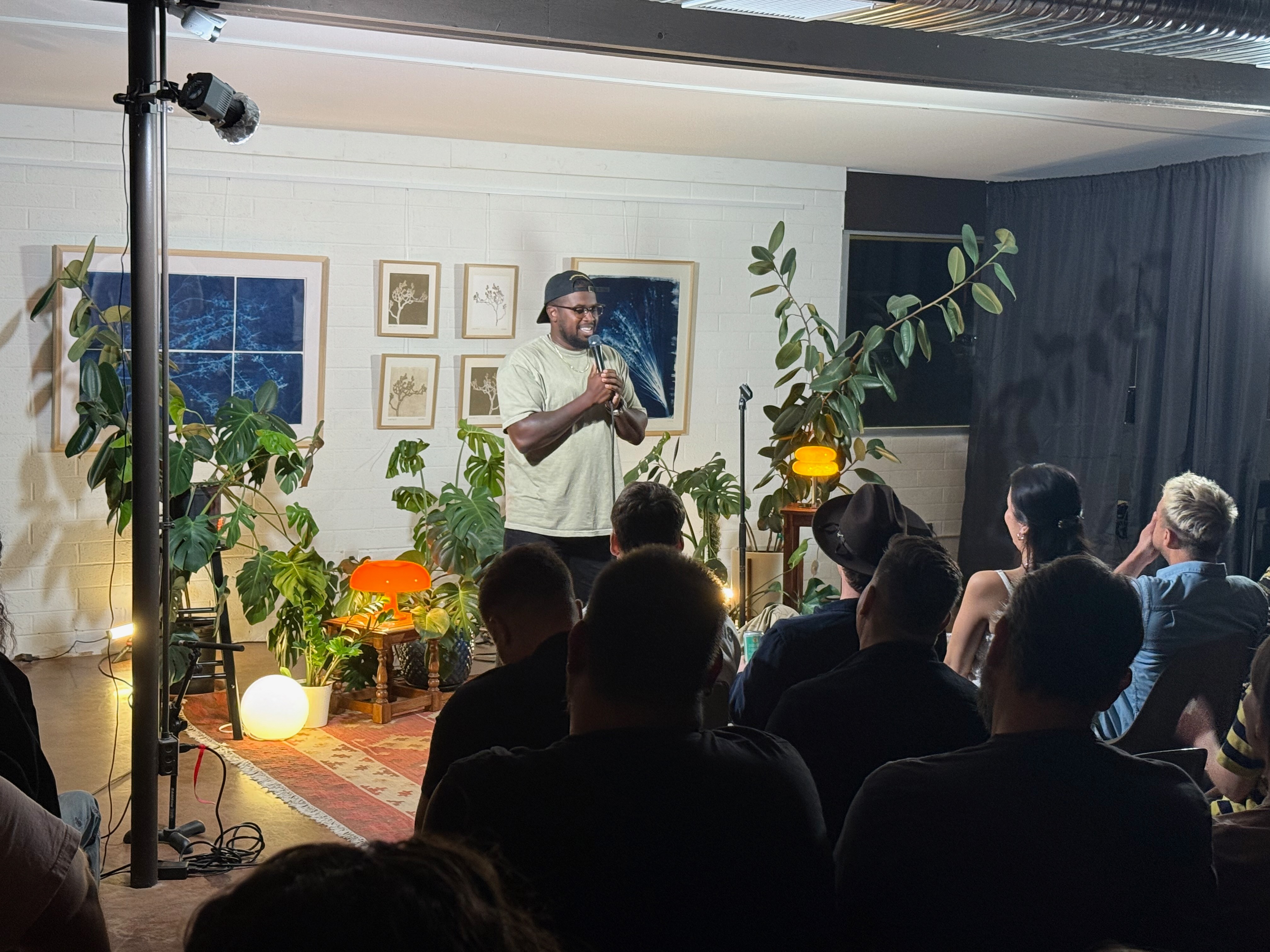 A man stands on a small stage with plants and framed pictures behind him, speaking to an audience in a cozy room.