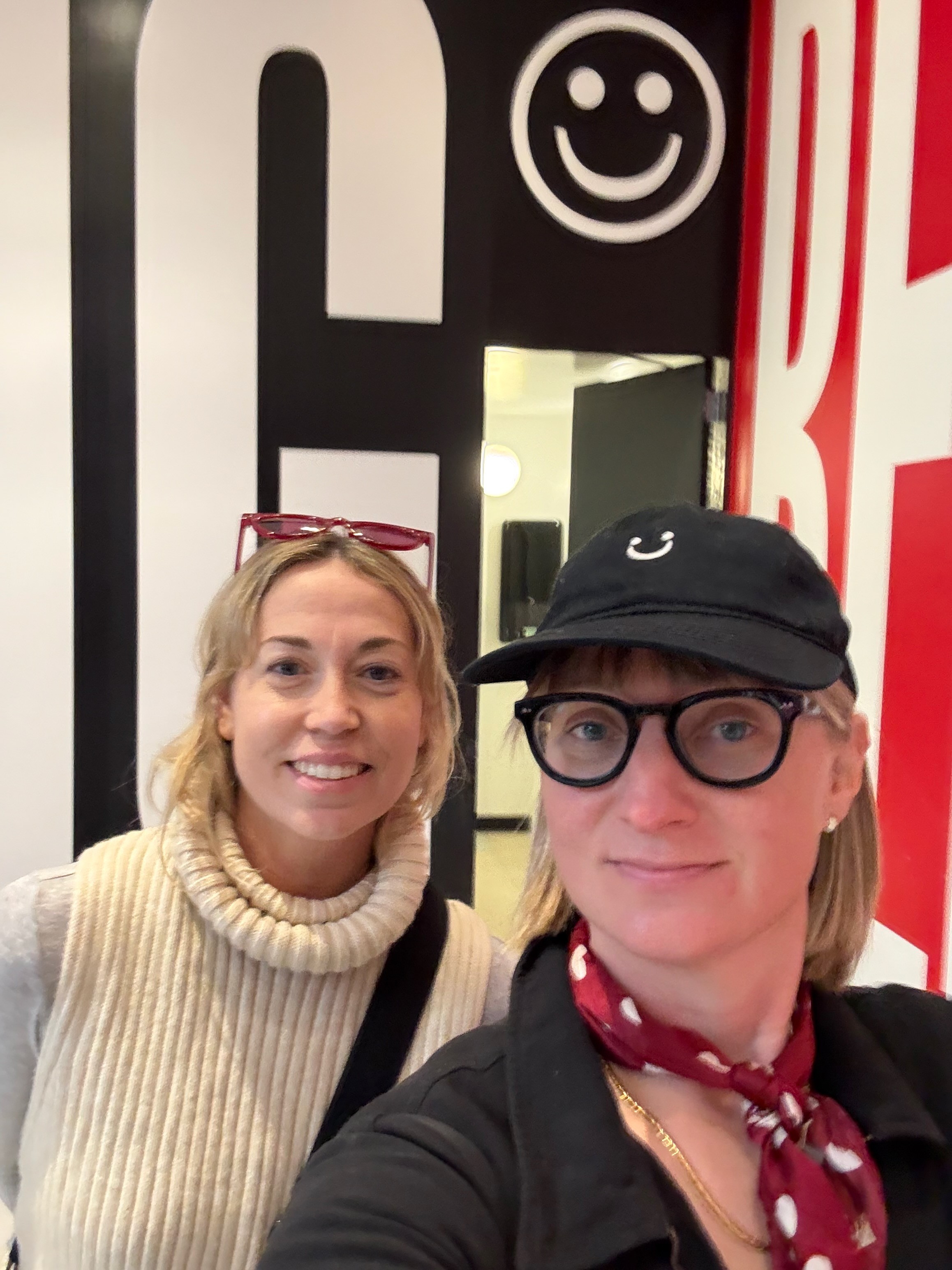 Two women smiling in front of a wall with a smiley face and large black and red letters.
