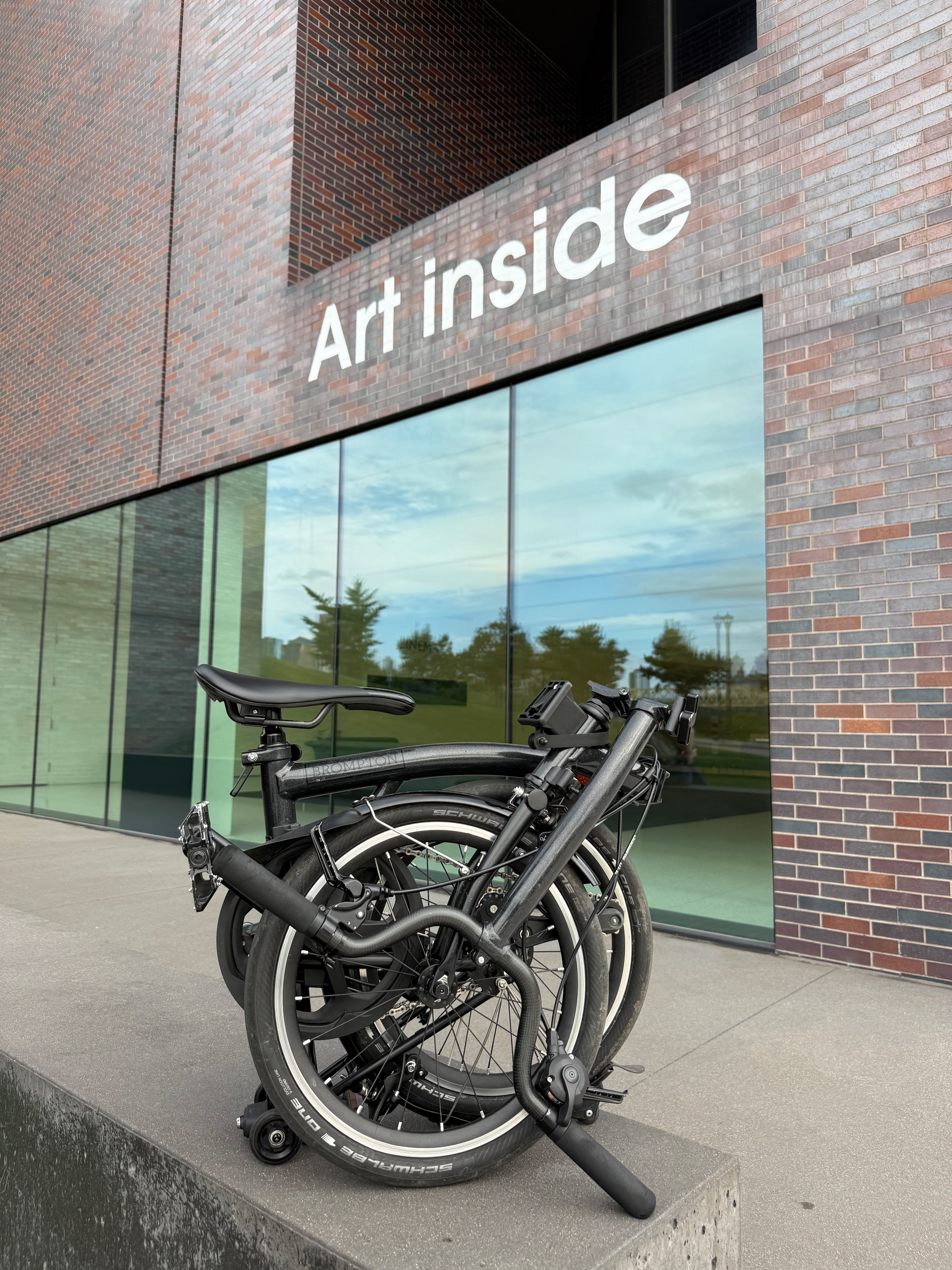 Folded black bicycle parked outside a building with large glass windows and a sign reading 'Art inside'.