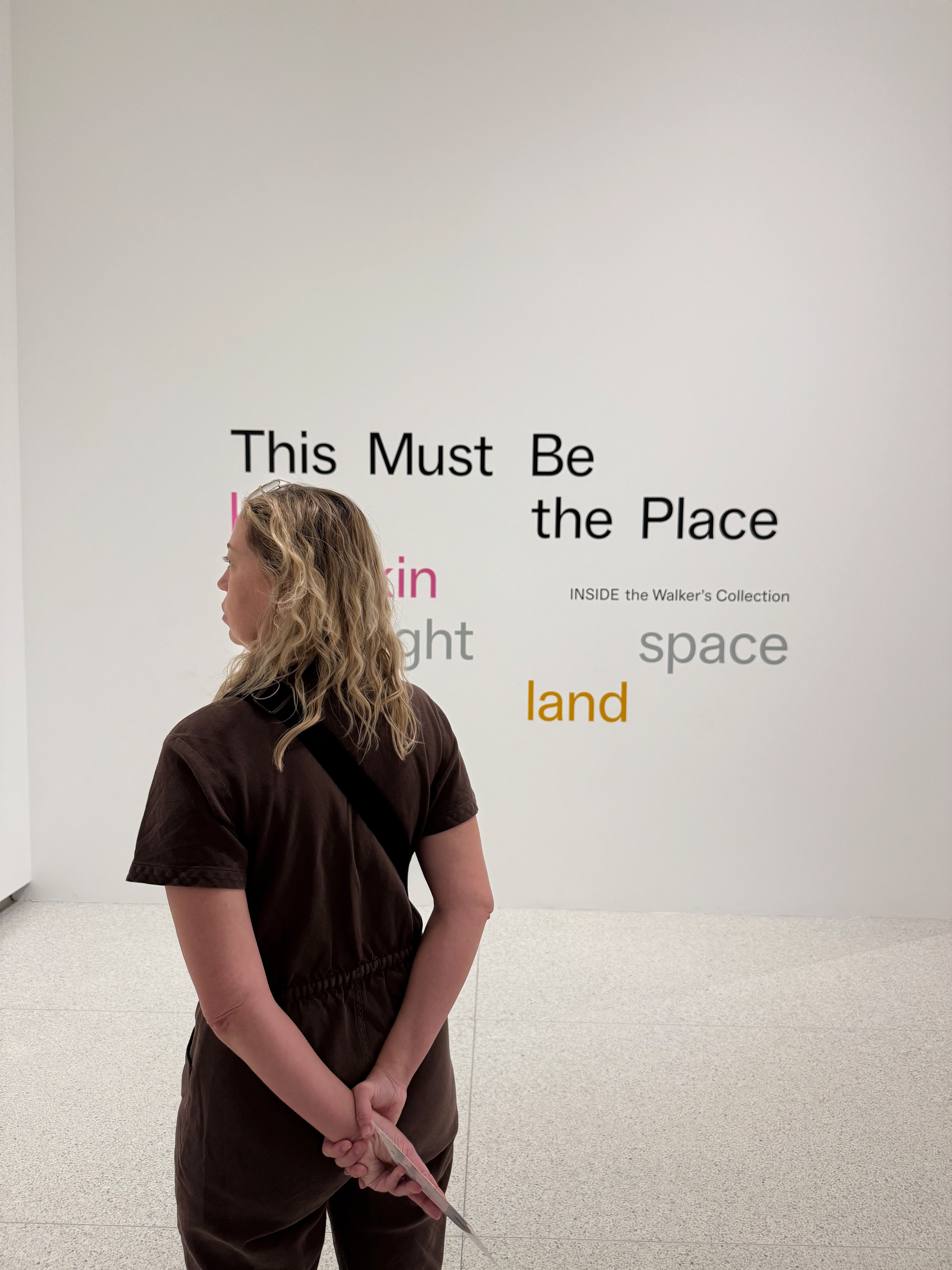 Person with curly hair stands in front of a wall with text, hands clasped behind back, facing away from the camera.